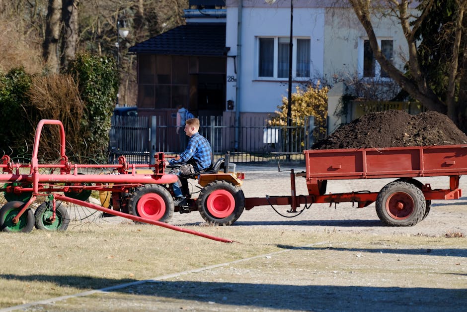 A young boy wearing a blue checkered shirt is seated on a small red tractor that is pulling a red trailer filled with dark soil, situated on a grassy area of a garden. The tractor has large red rear w