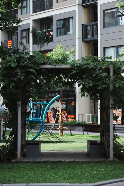 A landscaped communal area in Grahame Park, featuring a wooden pergola structure covered with lush green foliage and leafy branches creating a shaded sitting area. In the background, there is a small 